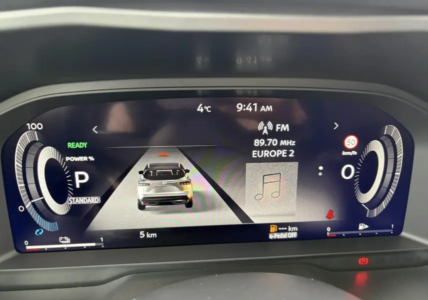 Affichage numérique du tableau de bord du Nissan Qashqai hybride, montrant la vitesse, la radio et l'icône du véhicule en vue arrière.