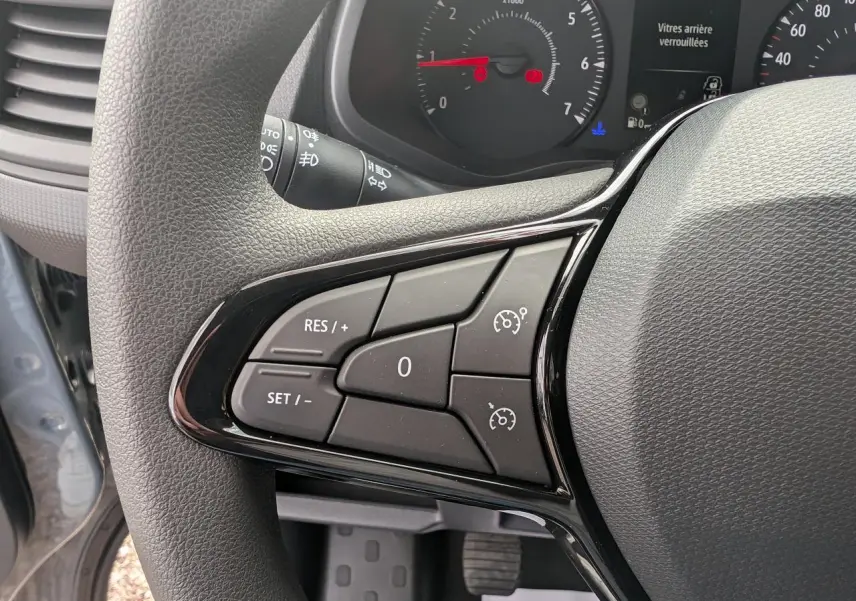 Gros plan sur les commandes de régulateur de vitesse au volant du Renault Trafic Fourgon gris, tableau de bord en arrière-plan.