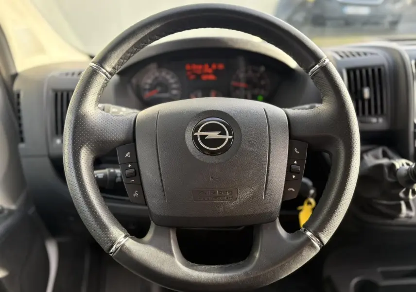 Gros plan sur le volant noir de l'Opel Movano Fourgon 2024 avec commandes intégrées et tableau de bord flou en arrière-plan.