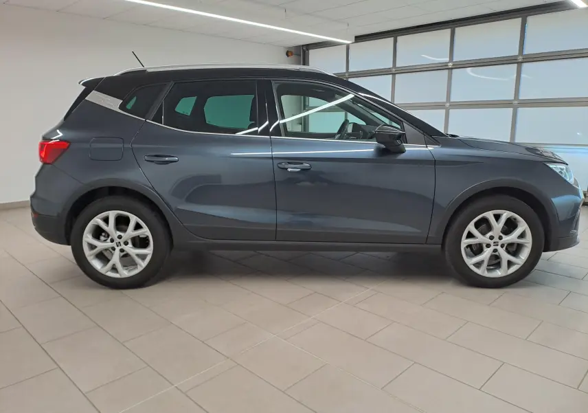 Vue de profil côté gauche d'un SEAT Arona 2024 gris magnétique avec toit noir dans un intérieur lumineux.