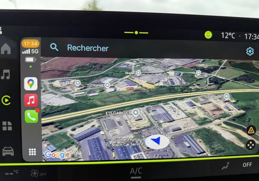 Écran de navigation GPS affichant une vue satellite en couleur avec itinéraire en bleu, dans un Dacia Duster gris Schiste.