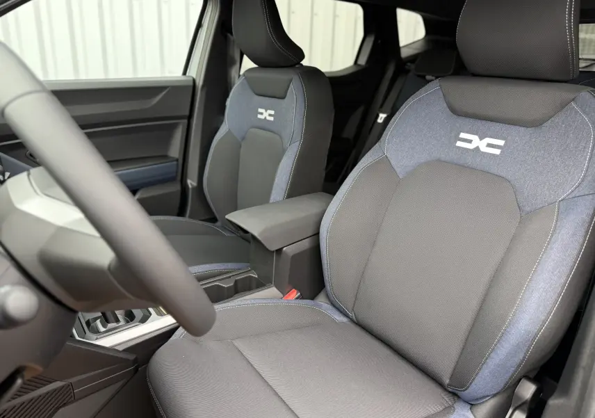 Vue intérieure du Dacia Duster 2025 montrant les sièges avant en tissu gris et bleu avec logo distinctif brodé.