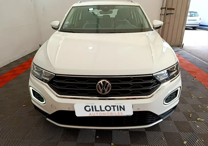 Vue frontale d'un Volkswagen T-Roc blanc avec feux LED allumés et calandre noire en intérieur garage.