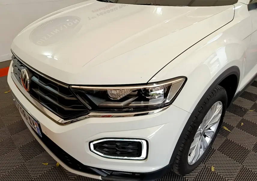 Vue 3/4 avant droit du Volkswagen T-Roc blanc, mettant en valeur ses phares LED et la calandre chromée.