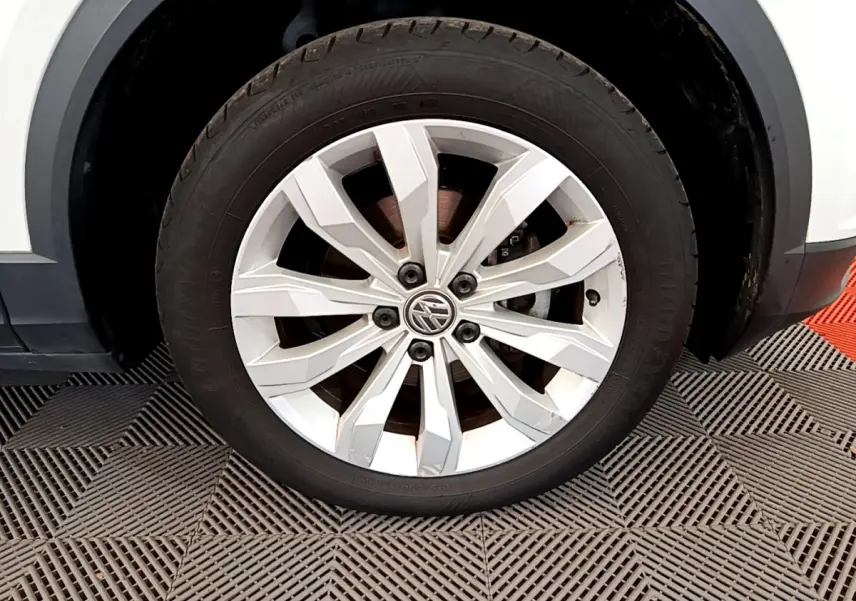 Gros plan sur la roue avant gauche avec jante argentée du Volkswagen T-Roc blanc, détail du pneu et logo VW visible.