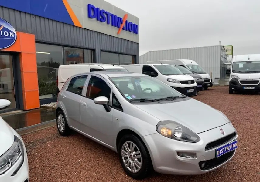 Fiat Grande Punto gris métallisé vue 3/4 avant droit avec phares allumés sur parking de concession.