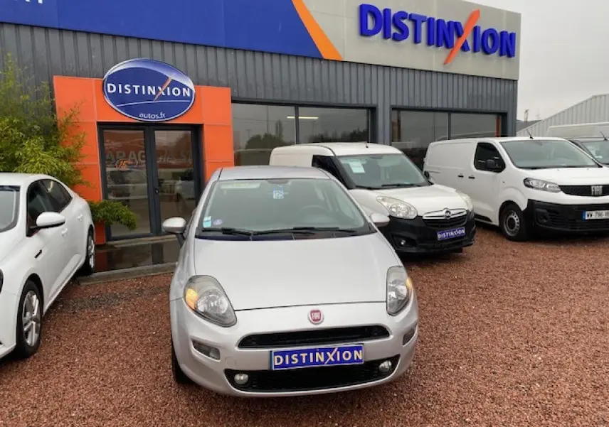 Vue avant d'une FIAT Grande Punto gris métallisé stationnée devant un showroom Distinxion par temps couvert.