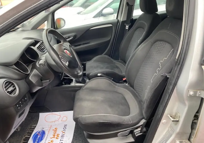 Vue intérieure côté conducteur de la Fiat Grande Punto 1.2i gris, montrant les sièges noirs et le volant multifonction.