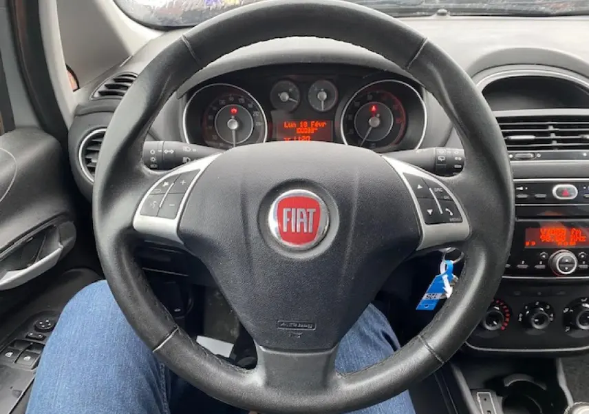 Vue intérieure centrée sur le volant noir de la Fiat Grande Punto 2015 avec commandes audio intégrées et tableau de bord visible.