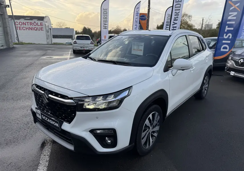 Suzuki S-Cross blanc 2023 en 3/4 avant droit, avec calandre noire et phares LED allumés sur parking.