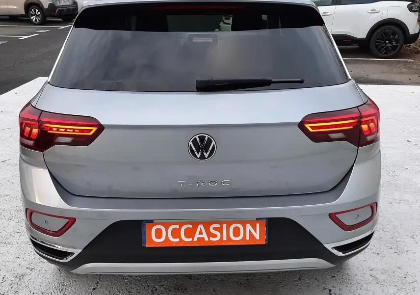 Volkswagen T-Roc gris pyrite vu en 3/4 arrière droit, feux arrière allumés sur parking extérieur.