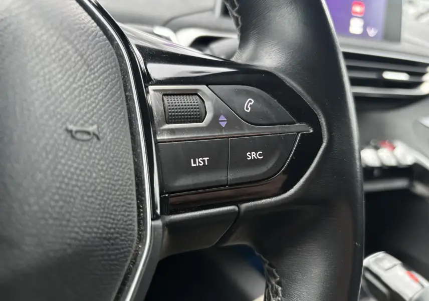 Gros plan sur les commandes gauche du volant noir du Peugeot 3008 blanc, avec écran tactile en arrière-plan.