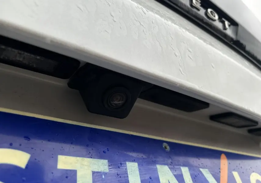 Gros plan sur la caméra de recul noire intégrée au hayon blanc nacré du Peugeot 3008, au-dessus de la plaque d'immatriculation.