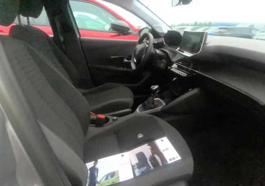 Vue intérieure côté conducteur de la Peugeot 208 gris clair, avec sièges tissu et écran tactile central visible.