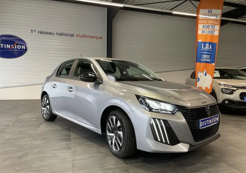 Peugeot 208 gris clair vue 3/4 avant droit en intérieur, avec calandre noire et feux LED distinctifs.