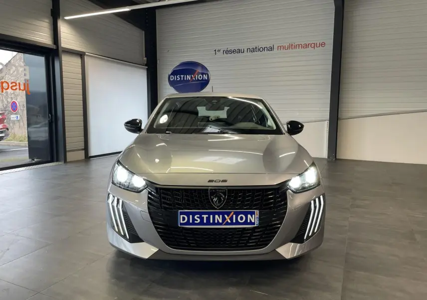 Vue frontale d'une Peugeot 208 gris clair avec calandre noire et feux LED à griffes allumés en intérieur showroom.