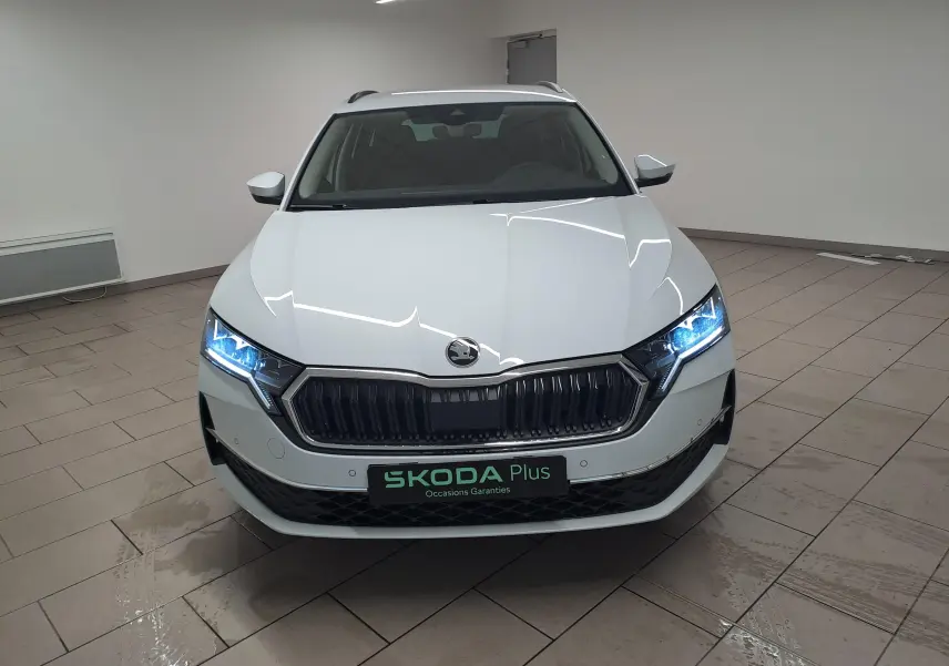 Vue frontale d'une Skoda Octavia Combi 2.0 TDI blanche Moon avec phares allumés dans un intérieur neutre.