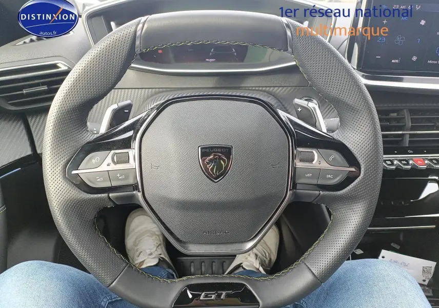 Vue rapprochée du volant cuir perforé avec logo Peugeot et badge GT, tableau de bord et écran tactile en arrière-plan.