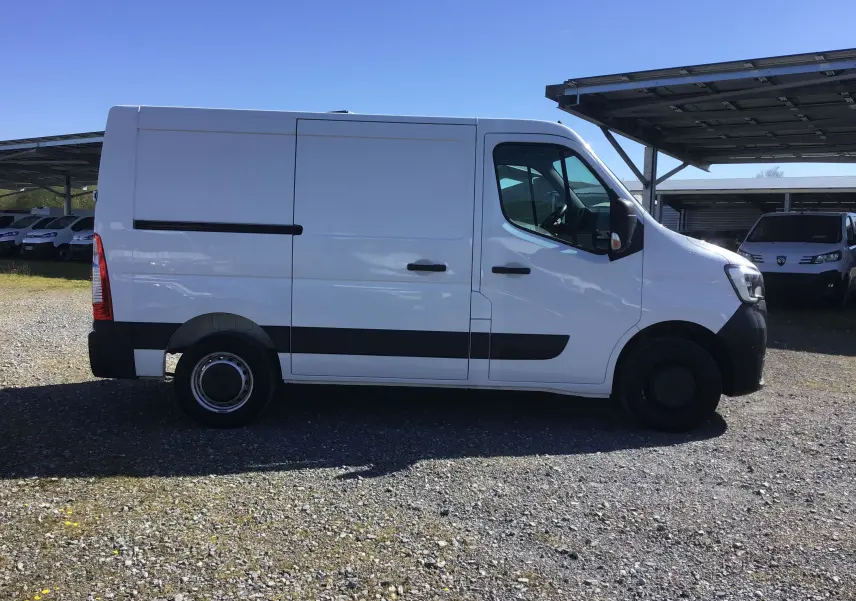 Vue de profil côté gauche d’un utilitaire Renault Master blanc avec bande noire et jantes acier, stationné sur un sol gravillonné.