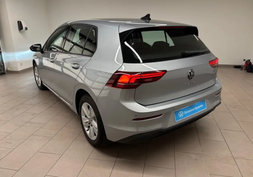 Vue 3/4 arrière droite d'une Volkswagen Golf 2025 argent reflet, avec feux arrière allumés dans un showroom.