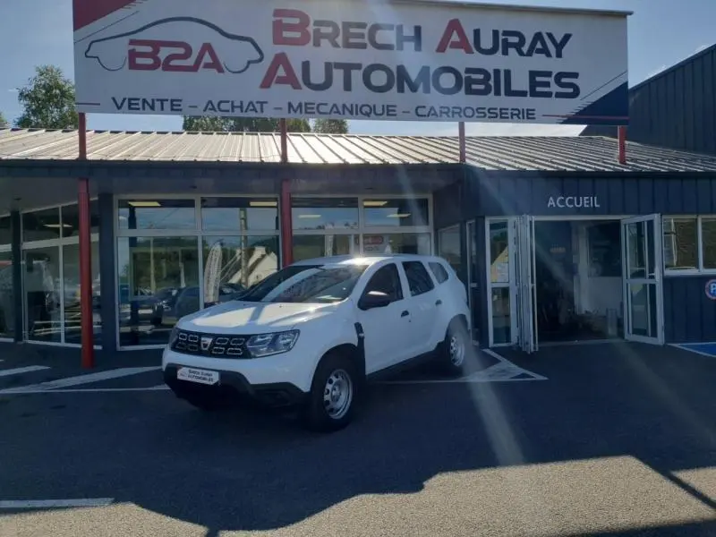Dacia Duster blanc en 3/4 avant droit stationné devant un garage Brech Auray Automobiles avec portes vitrées ouvertes.