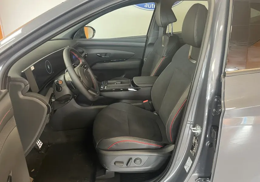 Vue intérieure centrée sur le volant noir avec surpiqûres rouges du Hyundai Tucson 2025, tableau de bord digital en arrière-plan.