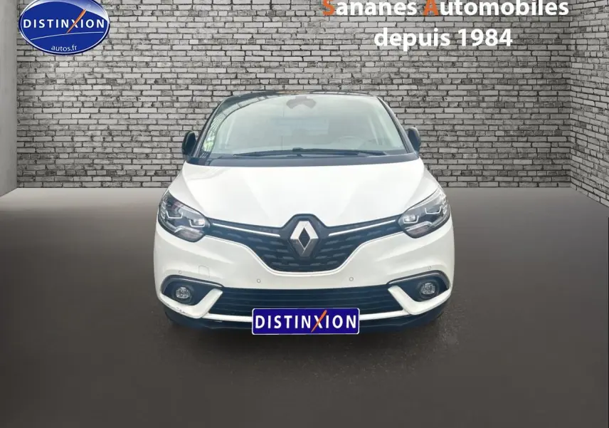 Vue avant d'un Renault Scénic blanc nacré avec calandre noire et phares LED, sur fond de mur en briques grises.