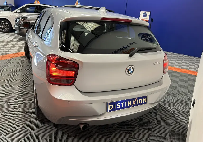 Vue arrière droite d'une BMW Série 1 gris clair 116d avec feux allumés dans un showroom.