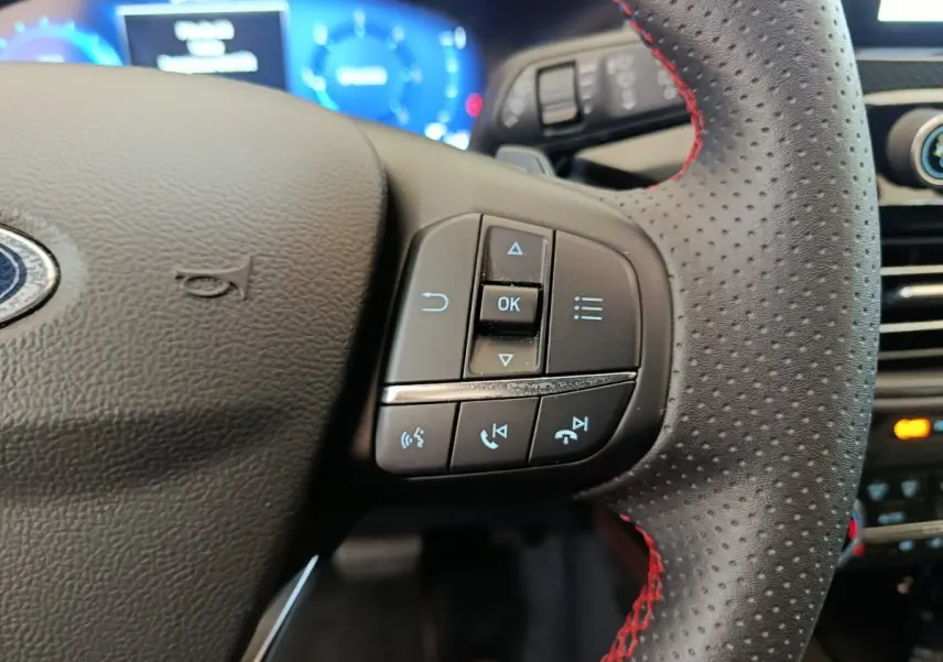 Gros plan sur le volant en cuir noir perforé avec surpiqûres rouges du Ford Kuga 2024, boutons multifonctions visibles.