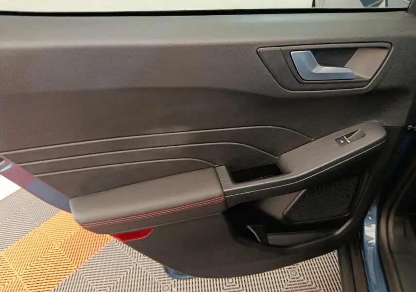 Gros plan sur la porte avant gauche du Ford Kuga bleu 2024, avec accoudoir noir et surpiqûres rouges.