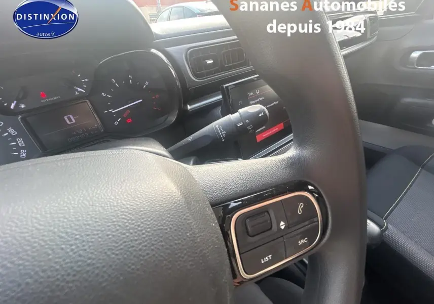 Vue intérieure centrée sur le volant et tableau de bord d'une Citroën C3 gris foncé avec commandes audio au volant.