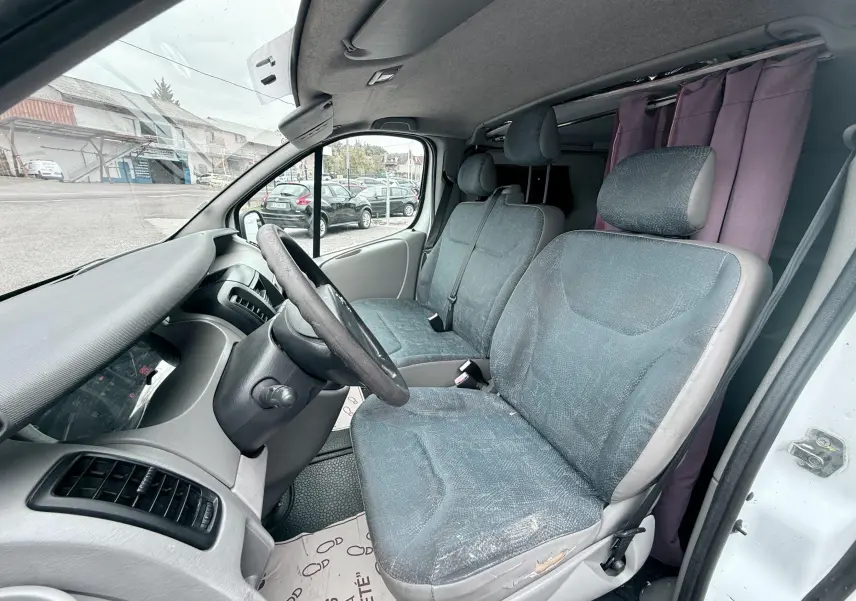 Vue intérieure côté conducteur du Nissan Primastar blanc de 2005, montrant les sièges en tissu gris bleu et le tableau de bord.