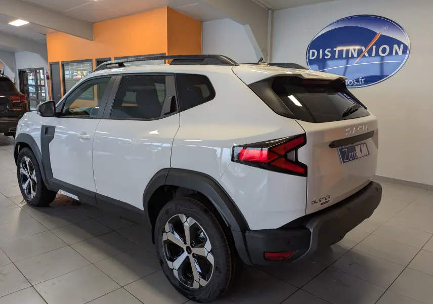 Vue 3/4 arrière droite du Dacia Duster blanc glacier 2025 en showroom, avec jantes alliage 18 pouces et feux arrière allumés.