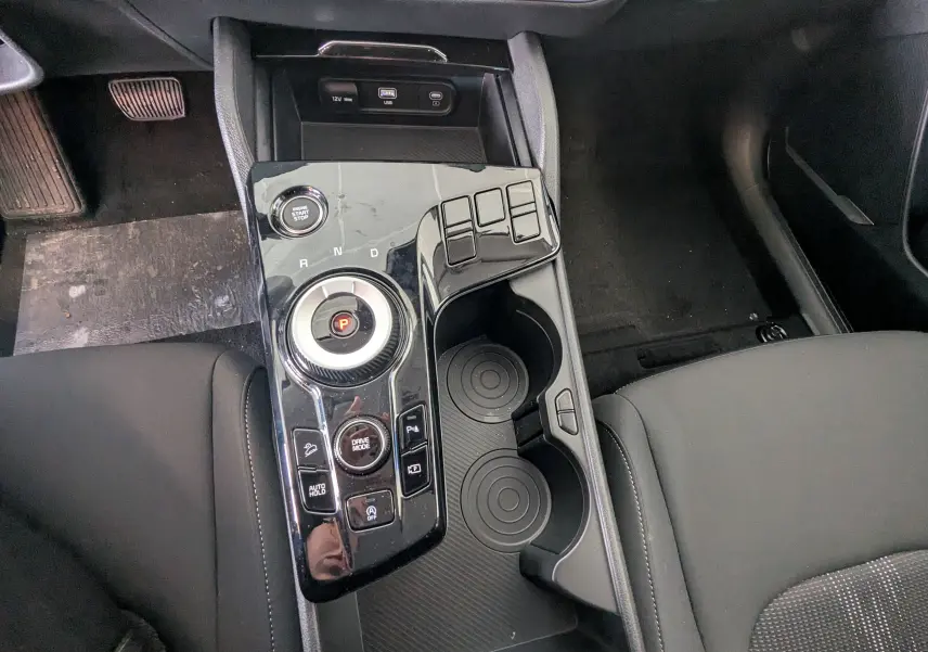 Vue plongeante sur la console centrale noire du Kia Sportage 2025, avec sélecteur rotatif de boîte auto et porte-gobelets.