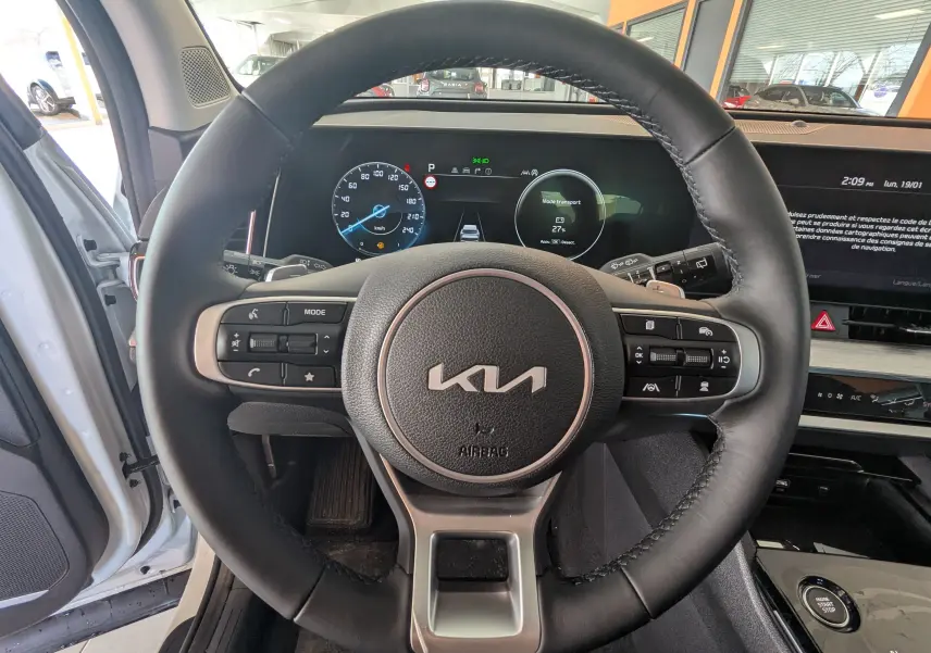 Vue intérieure centrée sur le volant noir du Kia Sportage blanc 2025 avec tableau de bord digital et écran tactile.