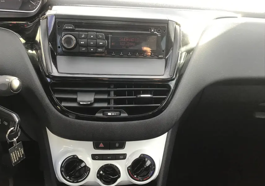 Vue rapprochée du tableau de bord noir et blanc de la Peugeot 208 1.2 PureTech 2018, avec radio et commandes de climatisation.