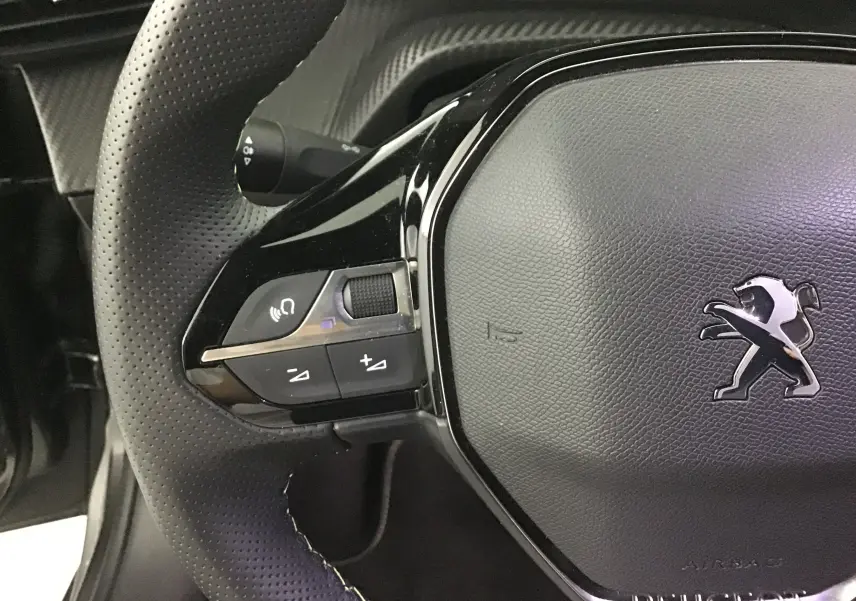 Gros plan sur le volant en cuir noir perforé du Peugeot 208 1.2 PureTech 100 GT, avec commandes intégrées et logo lion argenté.