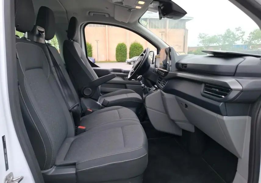 Vue intérieure côté conducteur du Ford Tourneo Custom blanc 2024, montrant sièges tissu gris et tableau de bord moderne.