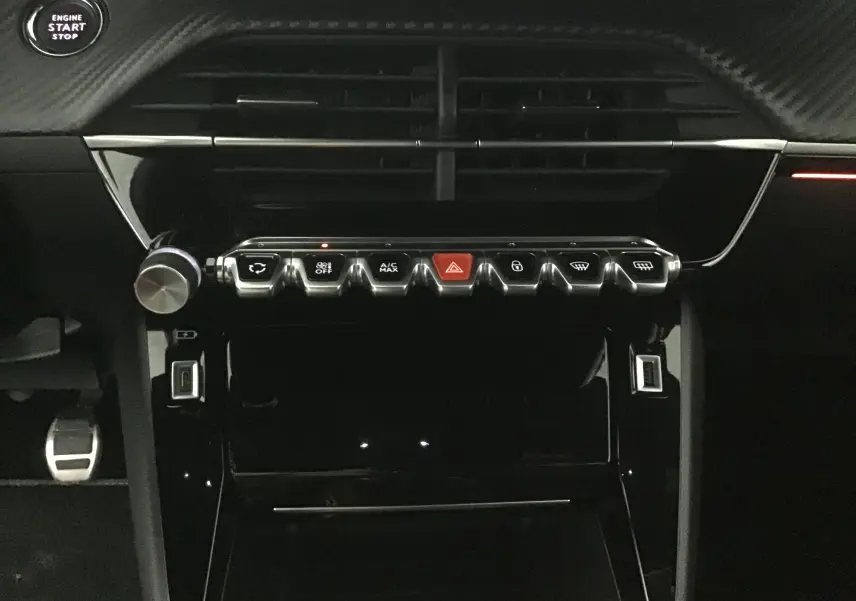 Tableau de bord intérieur de Peugeot 208 2023, vue frontale centrée sur les commandes et boutons chromés avec bouton d'alerte rouge.