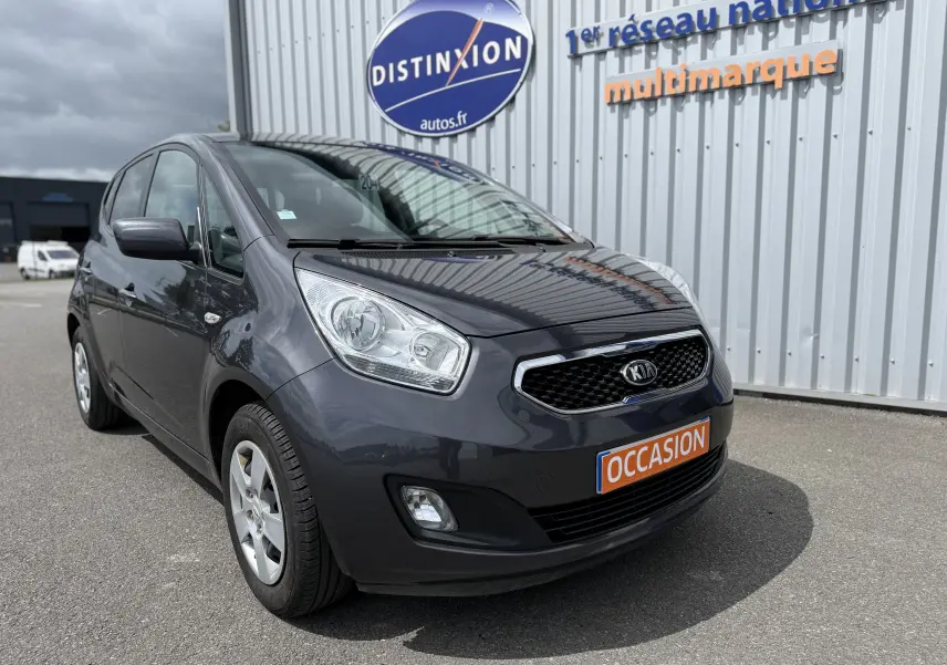 KIA Venga gris vue 3/4 avant droit, stationnée devant un bâtiment avec panneau Distinxion et plaque occasion.