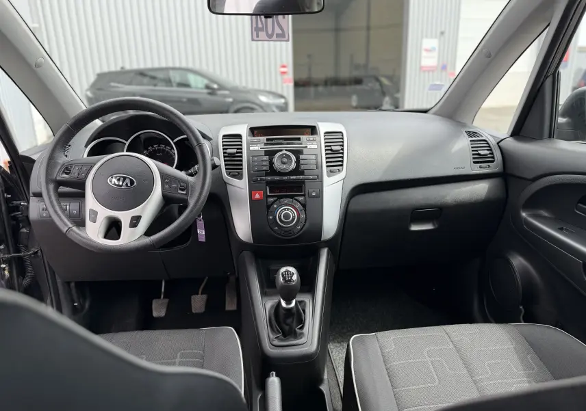 Vue intérieure avant de la Kia Venga 1.4 i 90 Active 2013, montrant le tableau de bord gris et la boîte manuelle.