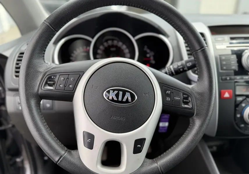 Gros plan sur le volant noir et gris du Kia Venga 1.4 i 90 Active de 2013, avec commandes intégrées et tableau de bord flou en arrière-plan.