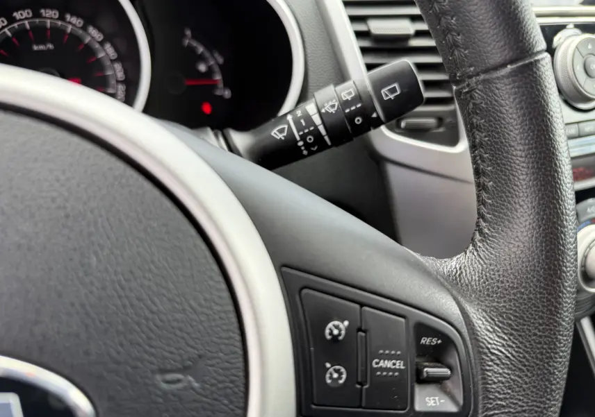 Gros plan sur le volant noir et le tableau de bord de la KIA VENGA 1.4 i 90 ACTIVE, avec commandes de régulateur de vitesse.