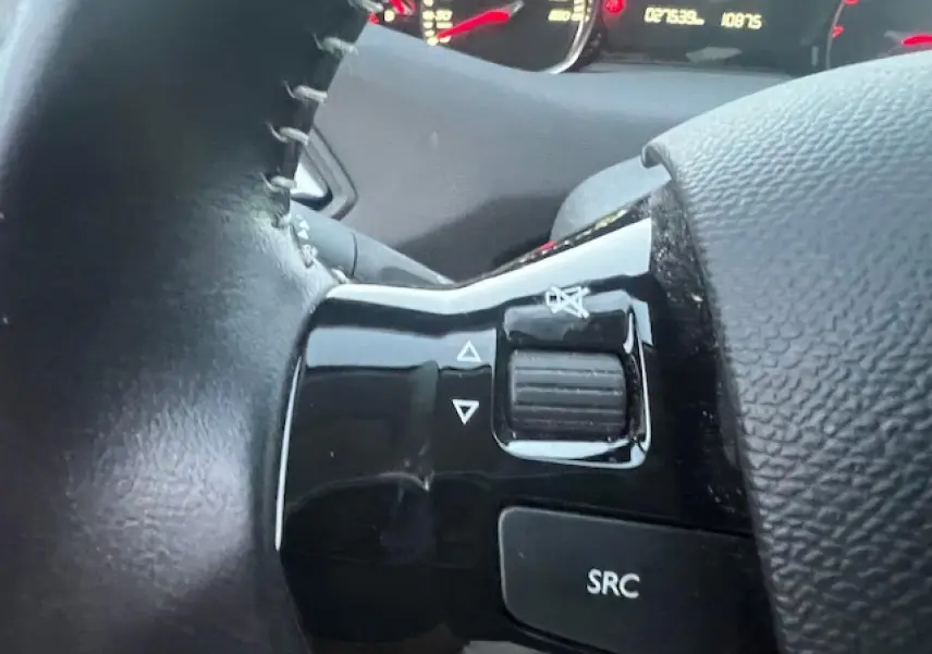Gros plan sur les commandes audio au volant noir de la Peugeot 308 blanche, avec tableau de bord en arrière-plan.