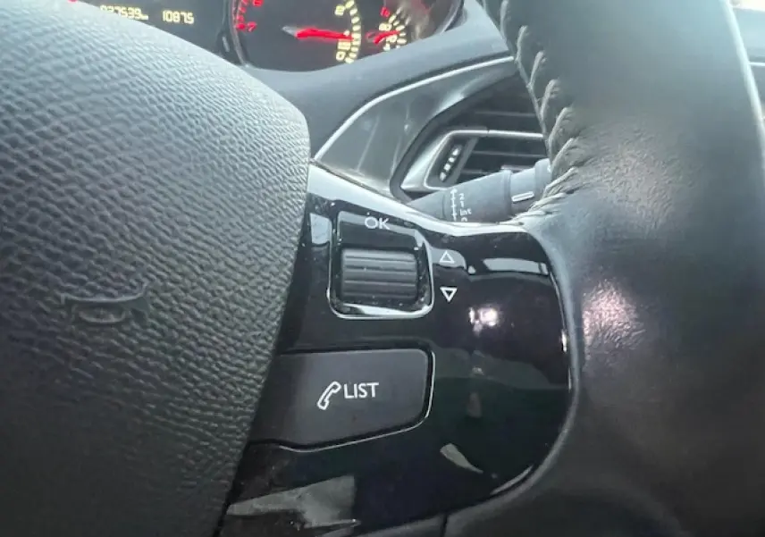 Gros plan sur les commandes au volant noir de la Peugeot 308 blanche, avec tableau de bord partiellement visible en arrière-plan.