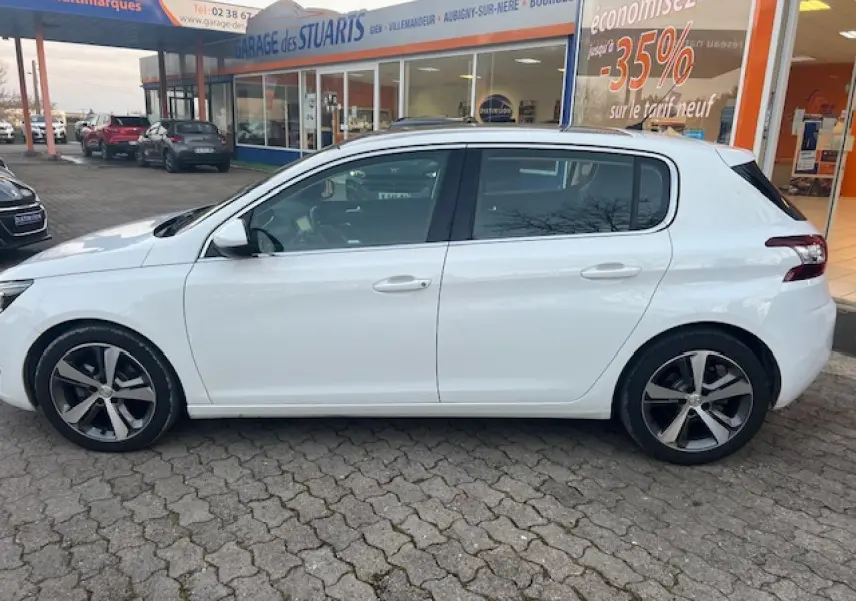 Peugeot 308 blanche vue de profil côté gauche, stationnée devant un garage avec toit panoramique visible.