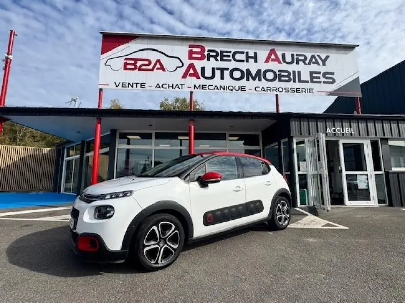Citroën C3 2017 blanc Banquise avec toit et rétroviseurs rouges, vue 3/4 avant droit devant un garage.