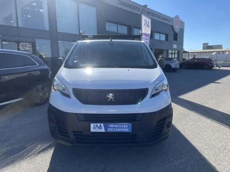 Vue frontale d'un Peugeot Expert Fourgon blanc 2023 avec calandre noire et logo lion visible, stationné devant un garage.