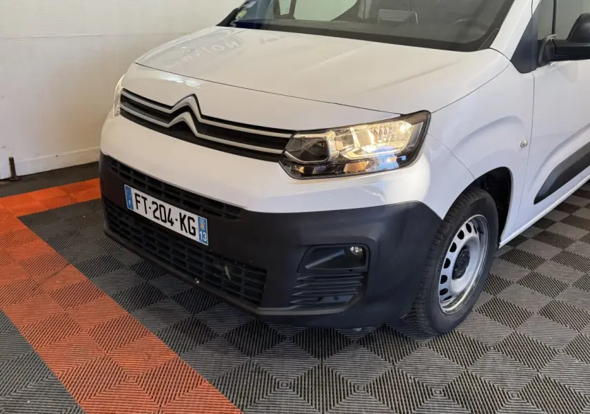 Vue 3/4 avant droit du Citroën Berlingo blanc avec pare-chocs noir et phares allumés sur sol carrelé.
