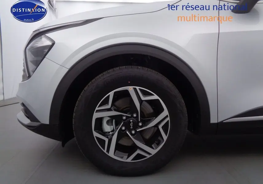 Gros plan sur la roue avant gauche et l'aile d'un KIA Sportage gris acier métal, avec jante bicolore et phares partiellement visibles.